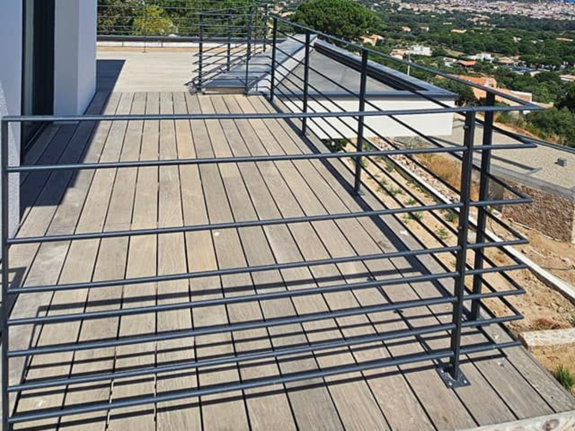 Garde-corps en métal noir sur une terrasse en bois surplombant une colline et des bâtiments lointains.