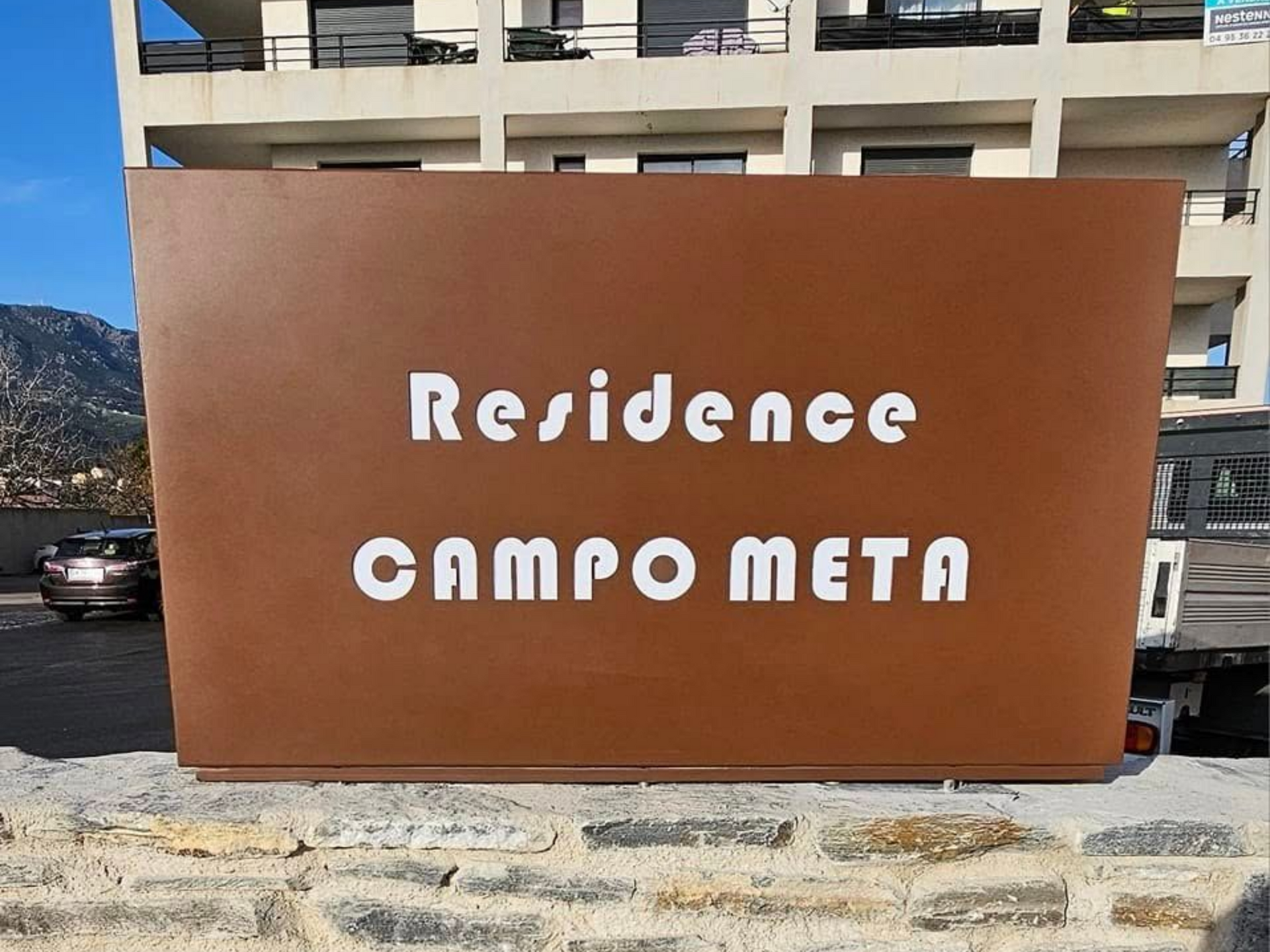 Panneau marron avec texte blanc indiquant Résidence CAMPO-META sur fond de bâtiment et de montagne.