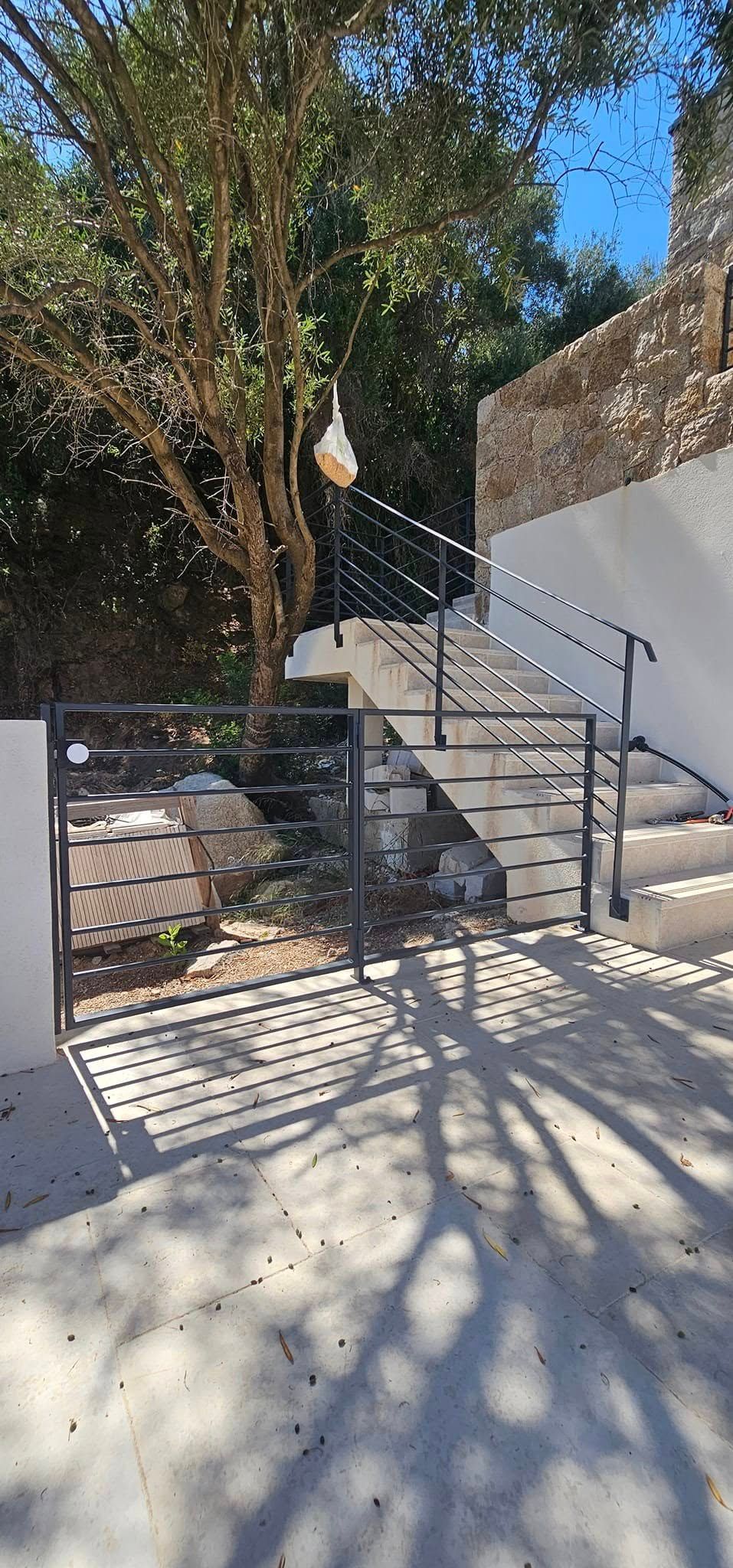 Un escalier en béton avec une rampe noire, menant à un bâtiment en pierre, ombragé par un grand arbre.