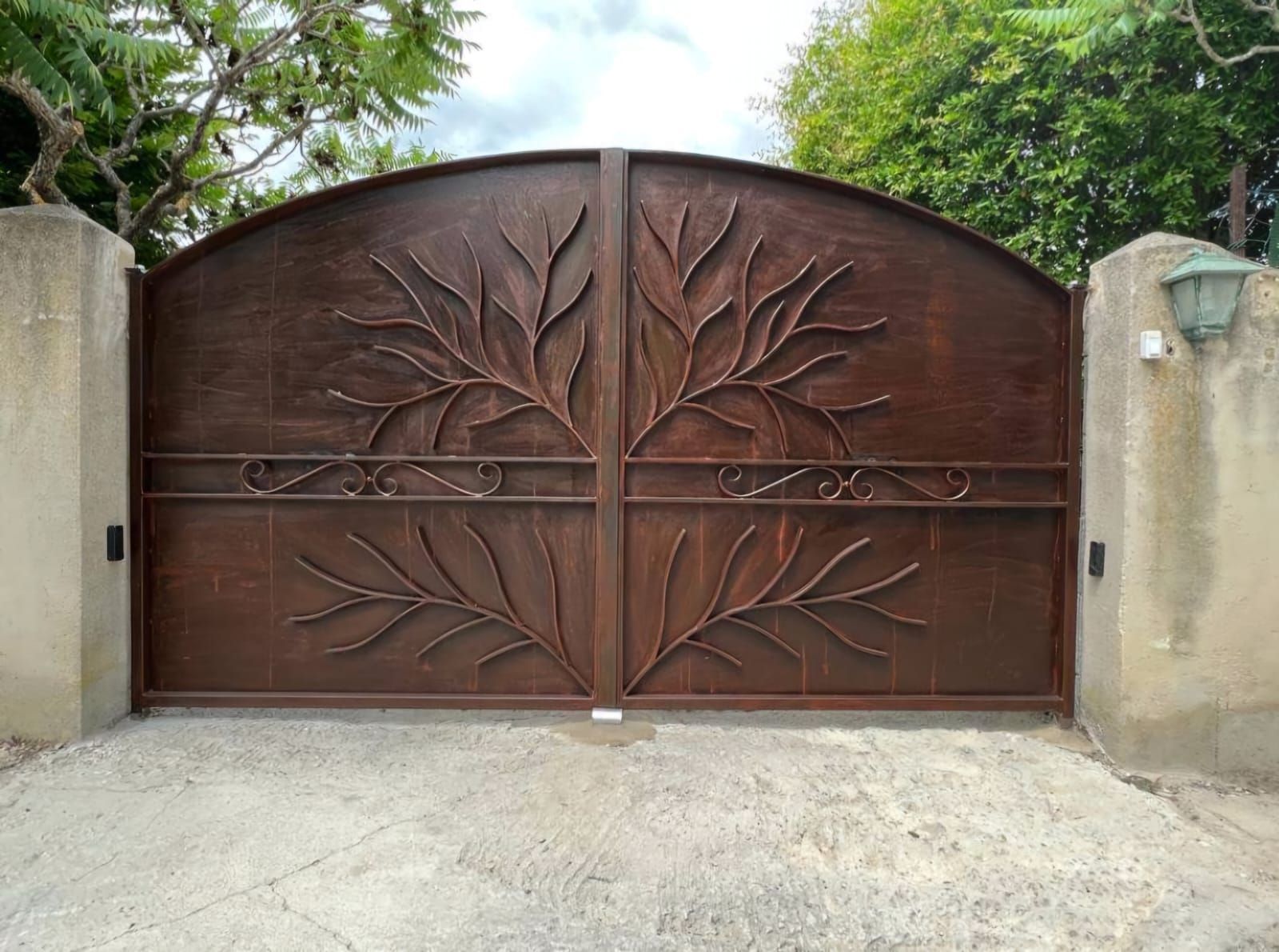 Portail en métal marron avec motif de branches d'arbres, entre des piliers en béton.
