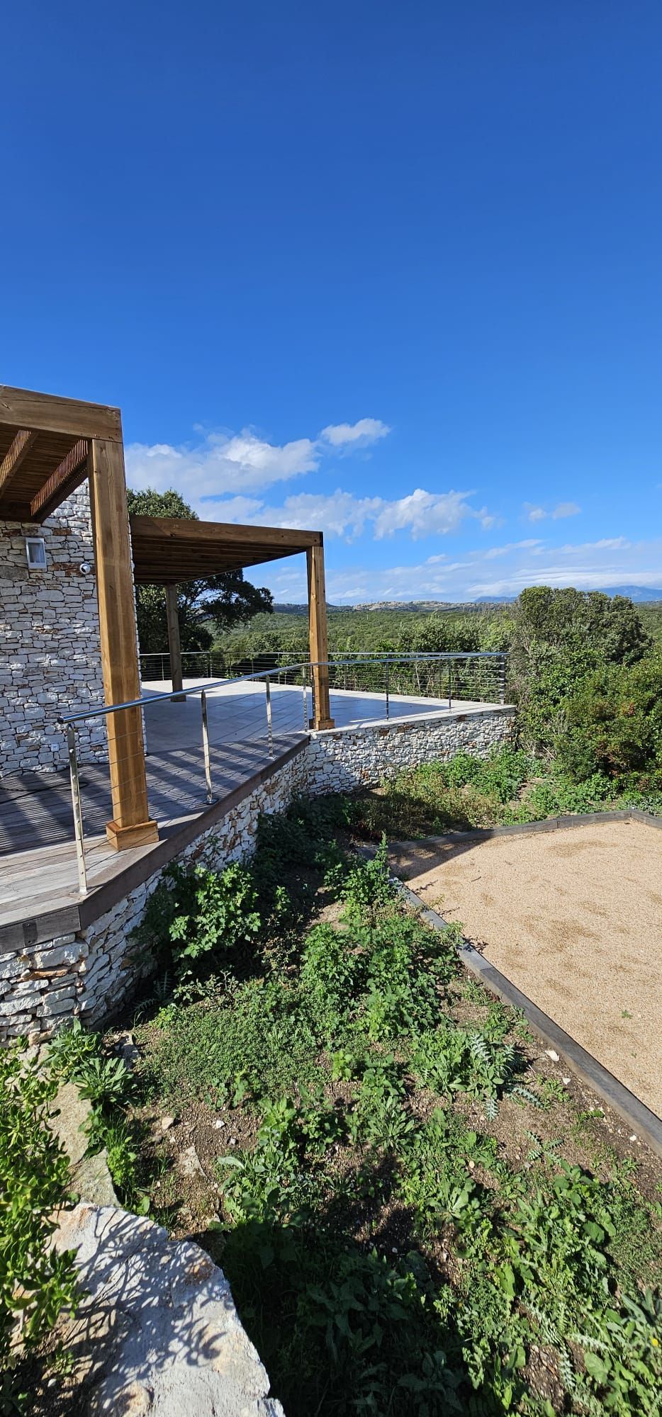 Garde-corps en métal noir sur une terrasse en bois surplombant une colline et des bâtiments lointains.