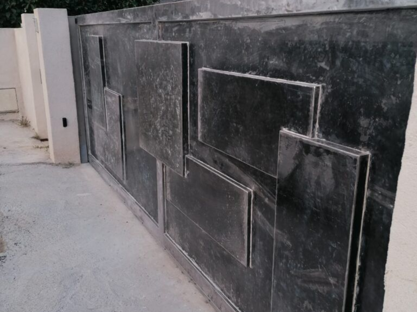 Portail en métal noir aux formes géométriques, à côté d'un pilier blanc. Sol en béton.