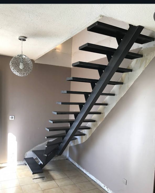 Escalier flottant en métal foncé contre un mur beige, avec des marches noires, dans un intérieur.