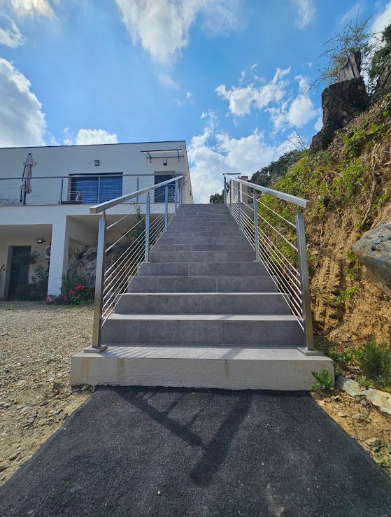 Escalier extérieur menant à un bâtiment blanc sous un ciel nuageux. Des rampes en acier inoxydable bordent les marches.