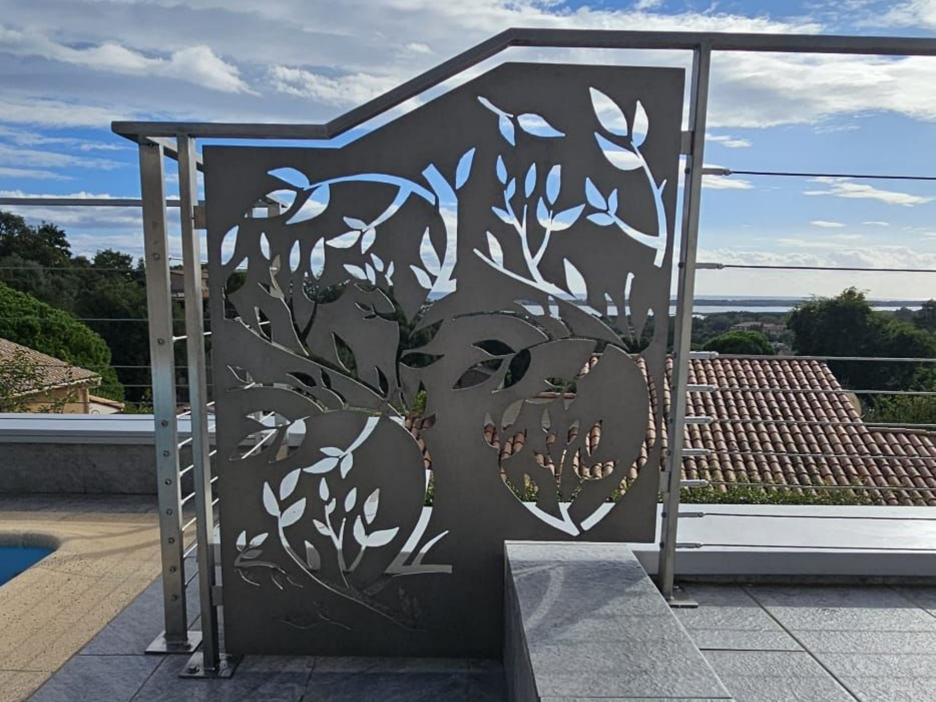 Panneau décoratif en métal à motif d'arbre sur un balcon, avec piscine et toits visibles.