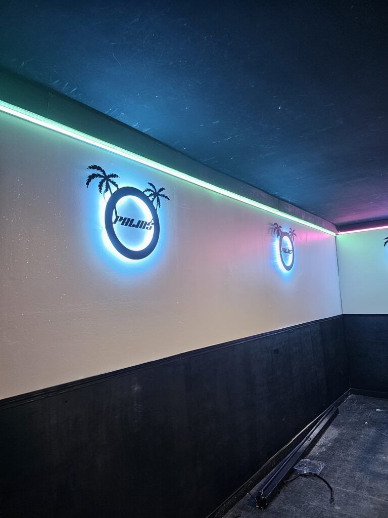 Une pièce aux murs noirs et blancs, avec un logo de palmier en métal sculpté et un éclairage à LED coloré.