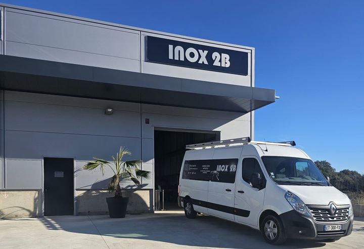 Camionnette blanche garée devant un atelier avec un panneau INOX 2B sous un ciel bleu.