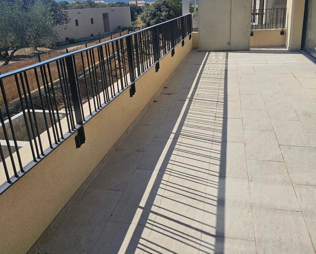 Balcon avec balustrade noire, sol en pierre et ombres projetées par la lumière du soleil.