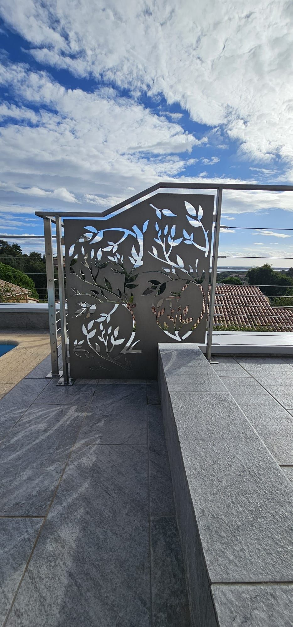 Portail décoratif en métal avec découpes de feuilles, sur un patio en pierre donnant sur un paysage sous un ciel bleu.