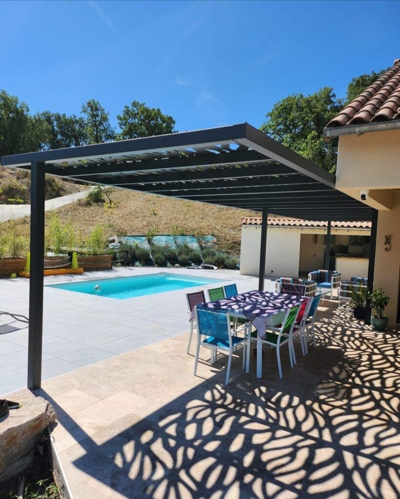 Pergola au-dessus d'une table à manger extérieure près d'une piscine, projetant des ombres de feuilles sur la terrasse. Journée ensoleillée.