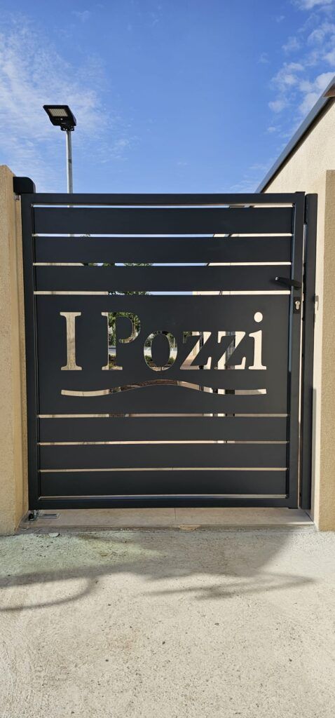 Portail en métal noir avec l'inscription I Pozzi découpée au centre. Il est doté de barreaux horizontaux et se dresse devant un ciel bleu.