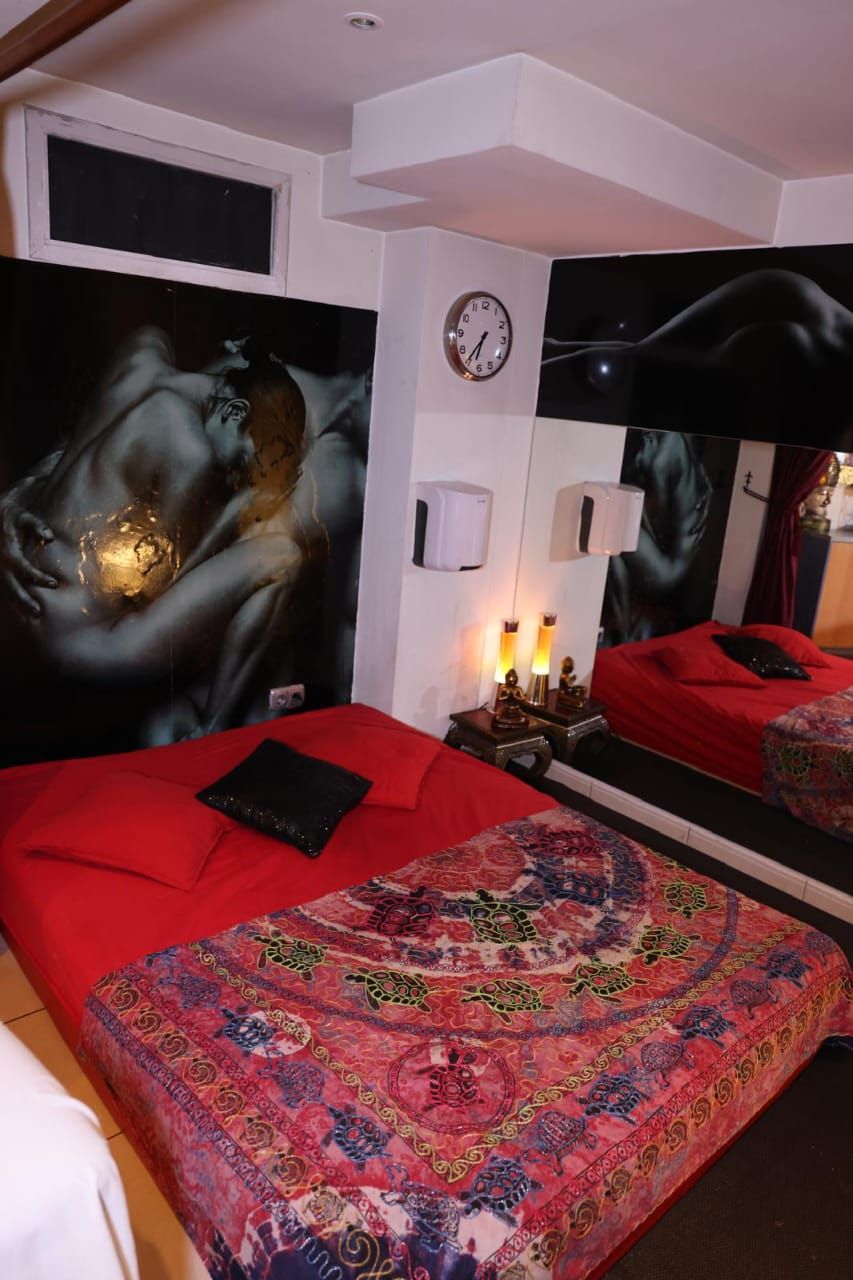 Dormitorio con colcha roja, cuadros coloridos en la pared, espejo y paredes blancas.