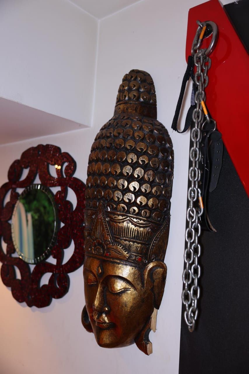 Máscara de pared dorada tallada con tocado ornamentado que cuelga junto a una pared roja y una decoración de cadena.