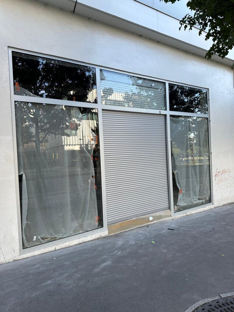 Vitrine avec de grandes baies vitrées et un rideau métallique gris, vue depuis le trottoir.