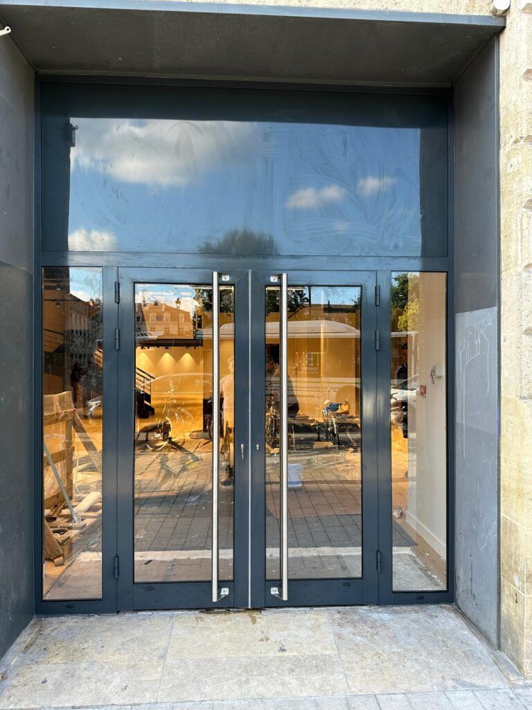 Entrée vitrée avec portes doubles et façade réfléchissante teintée de bleu sous un auvent en béton.
