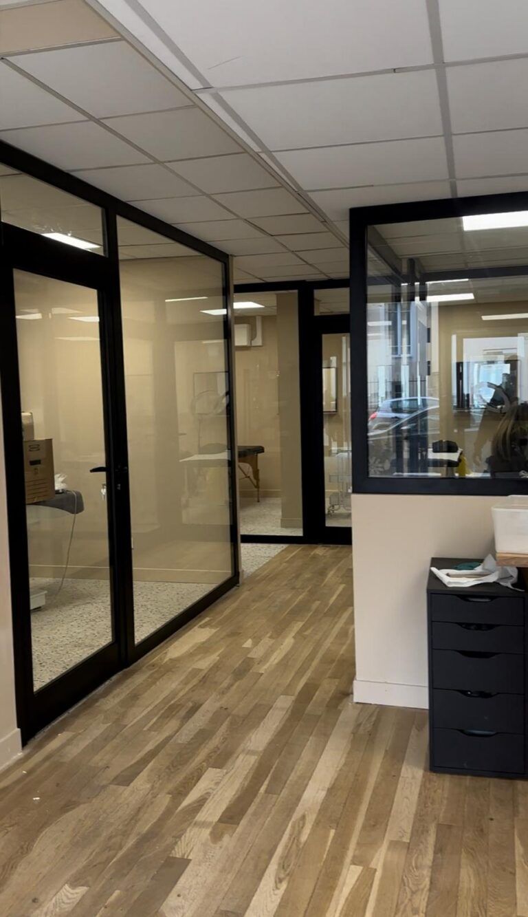 Couloir de bureaux avec des salles aux murs vitrés, un parquet en bois et des portes à cadre noir.