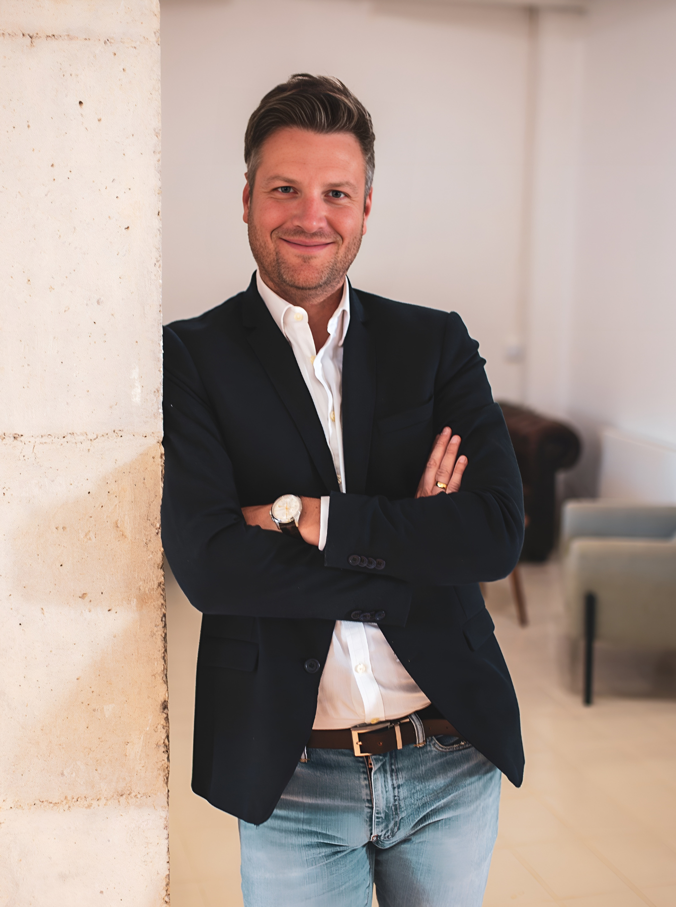 Ein Mann in Anzug und Jeans steht mit verschränkten Armen in einem Raum.