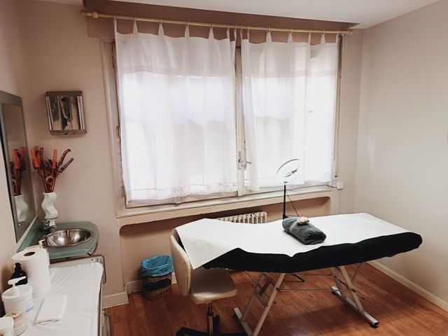 Sala de spa con mesa de masajes, lavabo y ventana con cortinas transparentes.