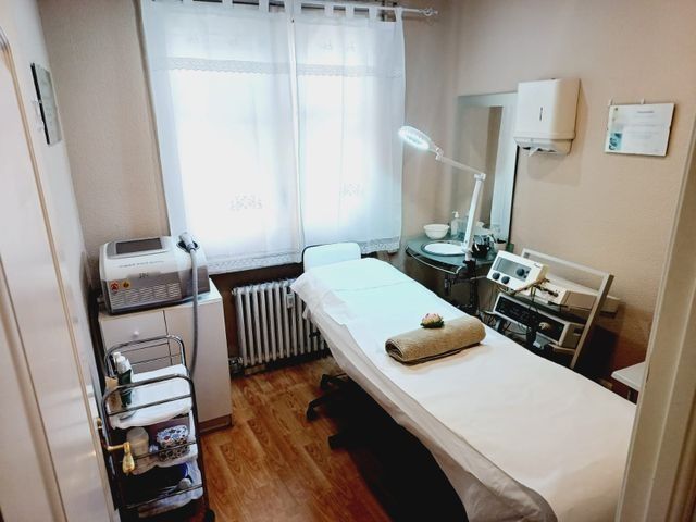 Una pequeña sala de tratamientos de spa con camilla de masaje, equipamiento y una ventana con cortinas transparentes.