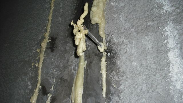 Un muro de hormigón con grietas verticales rellenas de espuma de poliuretano proyectada, endurecida y de color amarillento, que incluye un orificio de inyección sobresaliente.