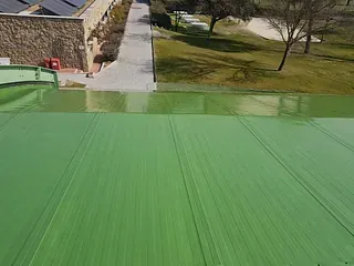 Vista desde un ángulo elevado de una superficie de techo lisa y de color verde brillante, junto a un edificio de piedra, una pasarela y una zona exterior cubierta de césped.