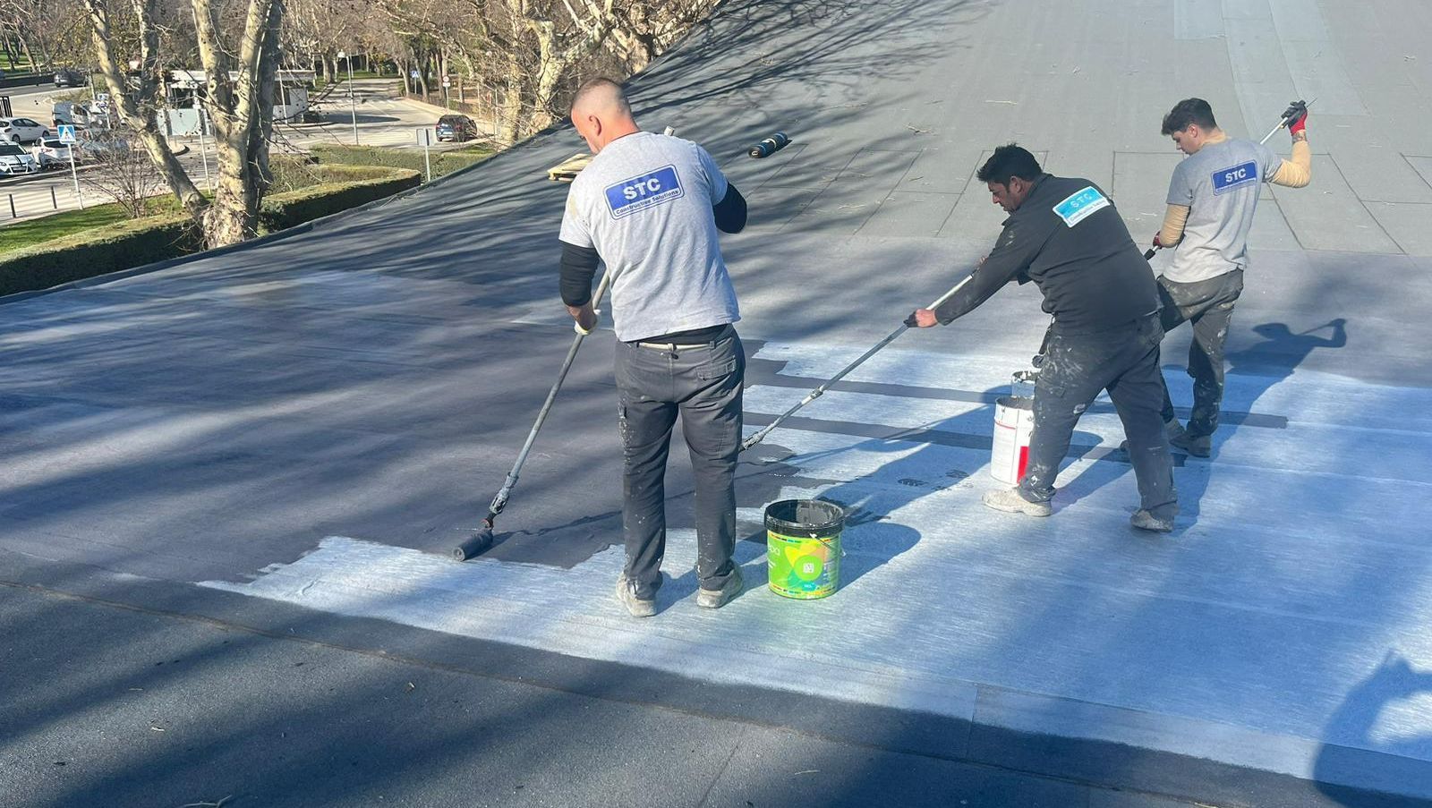 Tres trabajadores con camisas grises pintan de blanco un tejado plano con rodillos y cubos en un día soleado.