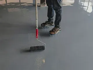 Un trabajador con zapatos de clavos utiliza un rodillo para extender una capa de resina epoxi gris sobre un suelo industrial liso.