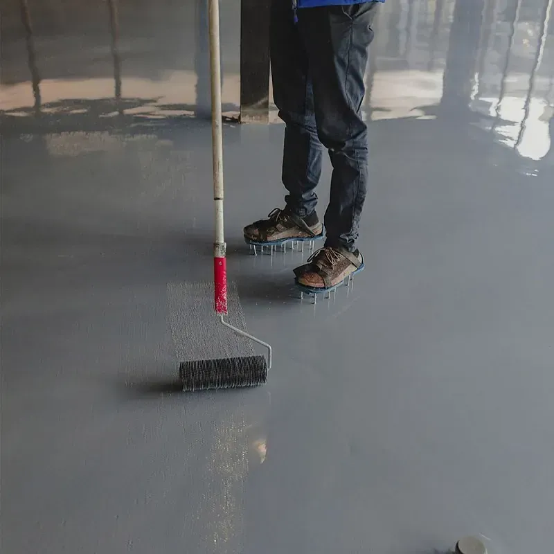 Una persona que lleva zapatos con clavos utiliza un rodillo para alisar la capa de epoxi gris recién aplicada en un suelo.