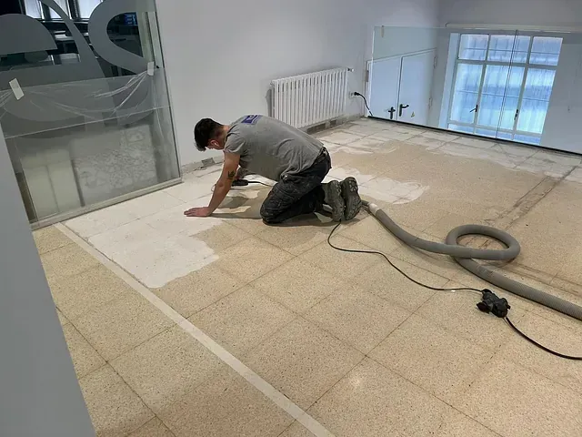 Una persona arrodillada sobre un suelo de baldosas, utilizando una herramienta conectada a una manguera para limpiar o decapar la superficie.