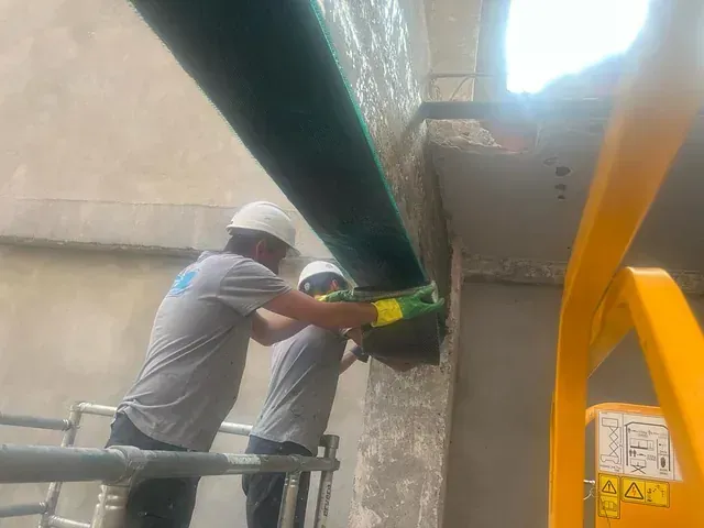Dos trabajadores con cascos y equipo de seguridad instalan una viga larga y oscura sobre una estructura de hormigón desde una plataforma elevadora.