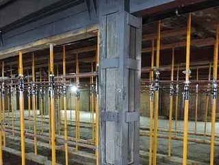 Un gran pilar gris reforzado se alza en una zona de construcción, sostenido por hileras de postes de apuntalamiento metálicos amarillos.