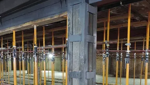 Una obra en construcción interior con una columna central de acero reforzado rodeada de numerosos postes de apuntalamiento amarillos que sostienen vigas.