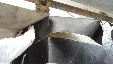 Trabajadores con trajes protectores blancos aplican una lámina de tejido de fibra de carbono negro a un elemento estructural.