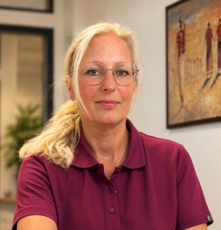 Porträt einer Frau in einem lila Poloshirt, die in einem Innenraum sitzt; im Hintergrund ist ein gerahmtes Kunstwerk zu sehen.