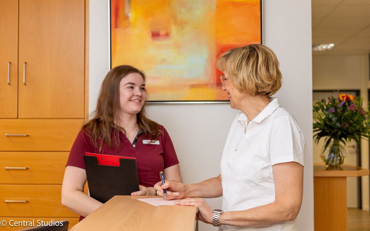 Eine Frau spricht mit einer anderen Frau am Tresen einer Hausarztpraxis für Allgemeinmedizin.