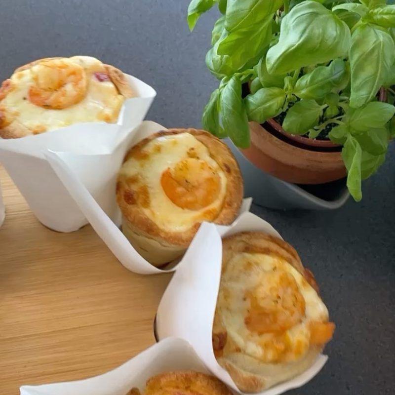Drie kleine hartige muffins in witte papiertjes op een houten plankje naast een basilicumplant.