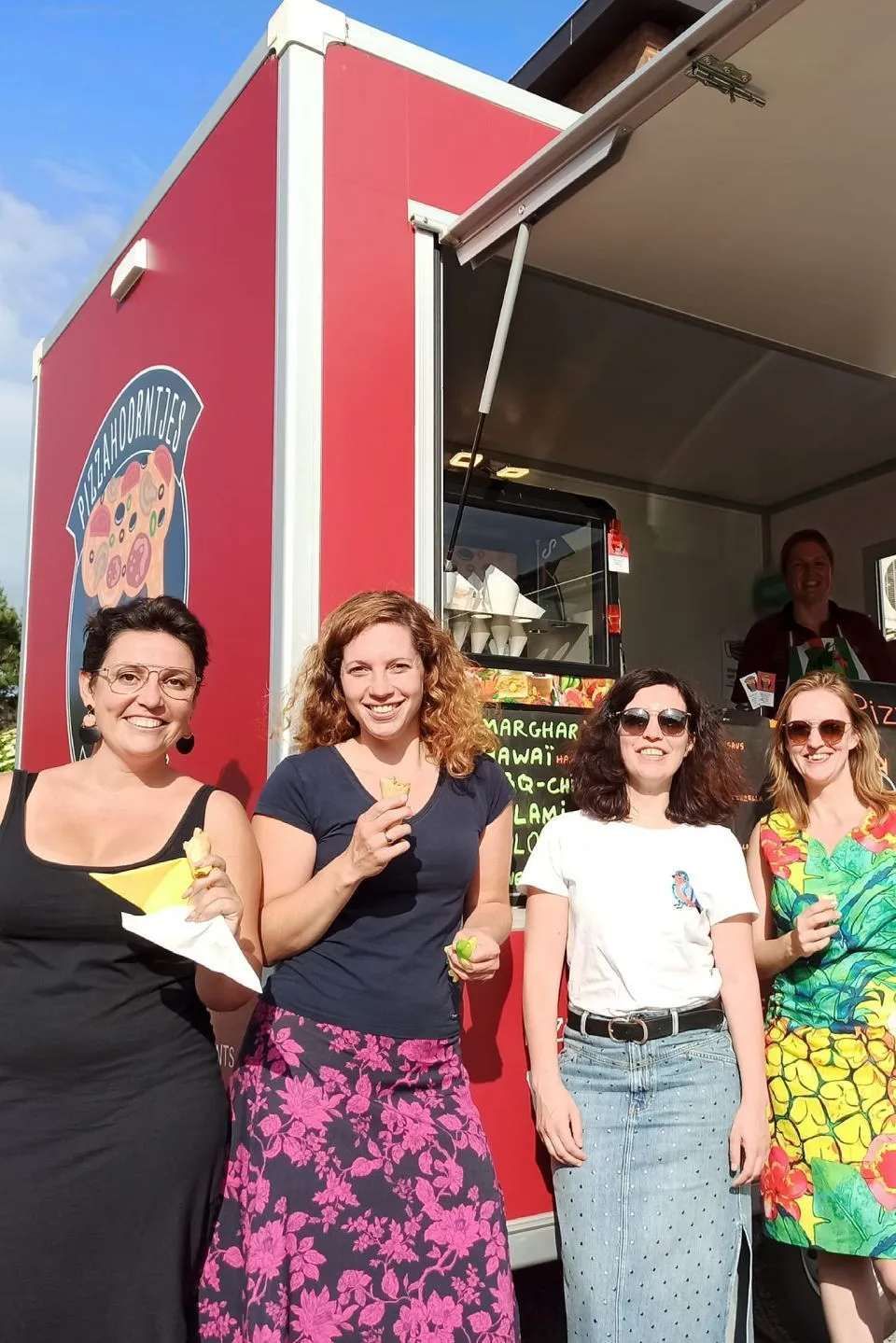 Vier vrouwen poseren voor een rode foodtruck, met drankjes in hun handen en een lachend gezicht.