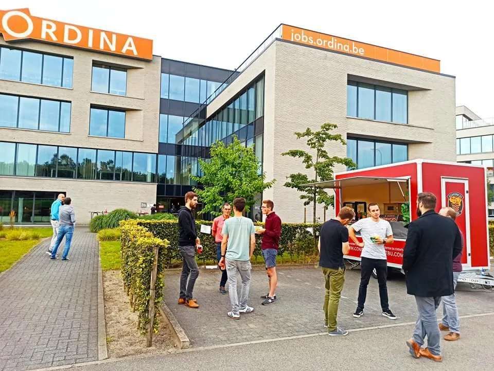 Mensen verzamelden zich op een bewolkte dag buiten een kantoorgebouw van Ordina, vlakbij een rode kiosk.