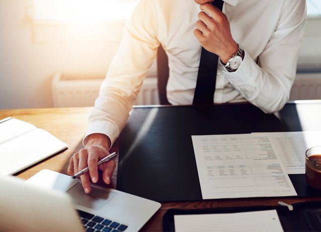 Hombre vestido formal, trabajando en un escritorio, con aspecto pensativo, con un bolígrafo en la mano, papeles y una computadora portátil presentes.