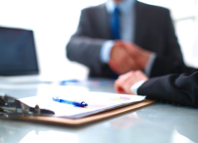 Dos hombres de negocios se dan la mano por encima de un portapapeles con un bolígrafo sobre un escritorio.