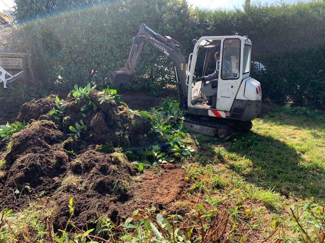 Une petite pelleteuse creuse dans un jardin, près d'une haie, déplaçant terre et végétation.