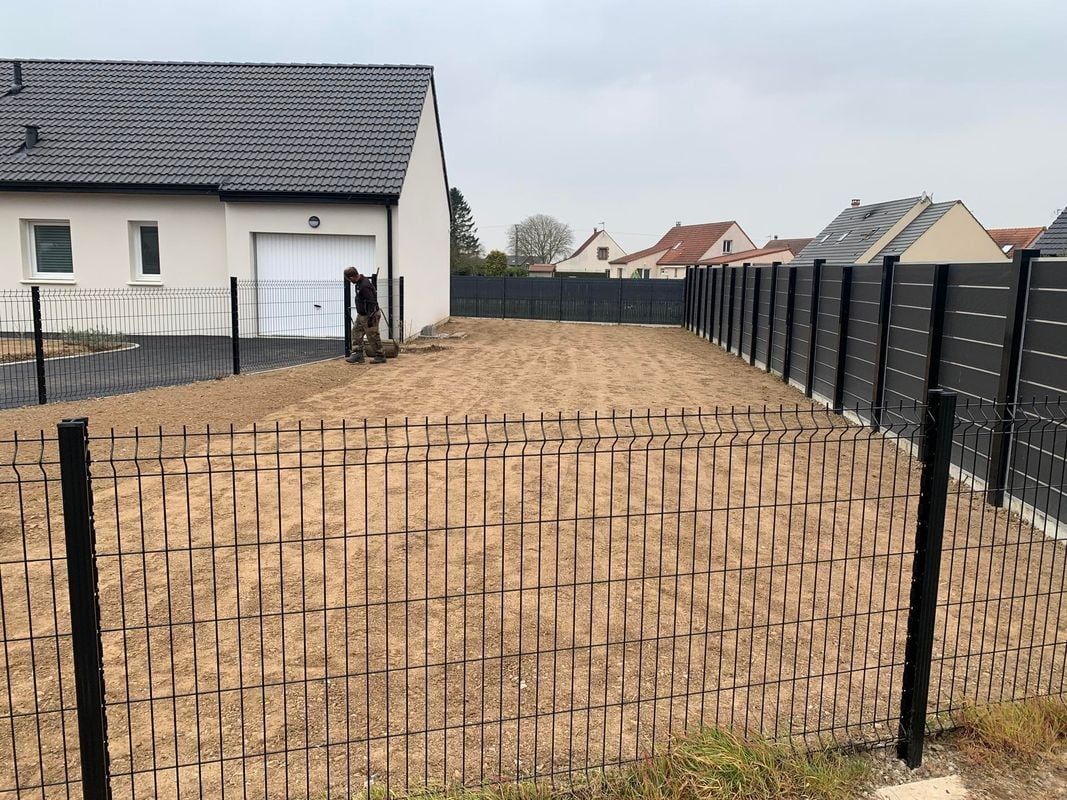 Cour en terre avec une clôture tressée devant la maison et une clôture noire sur le côté.