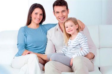 Familia sonriente sentada en un sofá blanco, vestida con ropa de colores claros en una habitación luminosa.