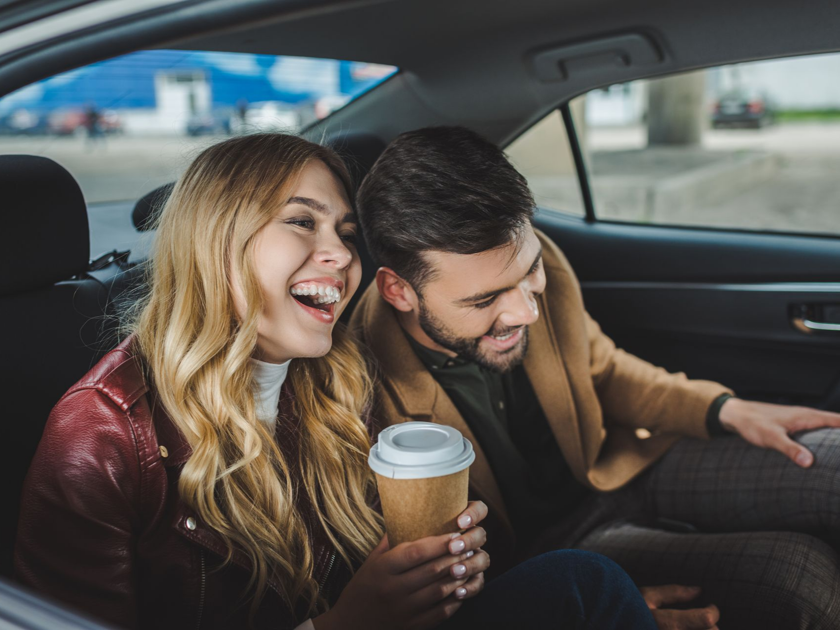 Un couple souriant dans une voiture ; la femme tient une tasse de café, l'homme se penche vers elle.