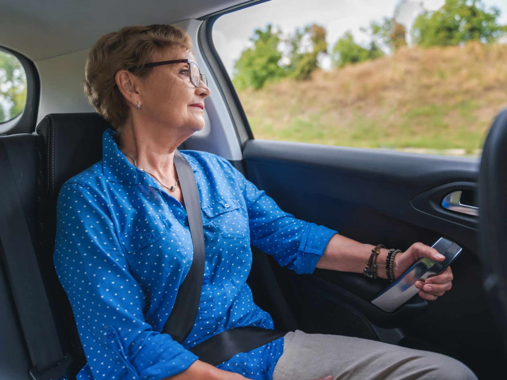 Une femme en chemise bleue, portant des lunettes et une ceinture de sécurité, regarde par la fenêtre de la voiture.