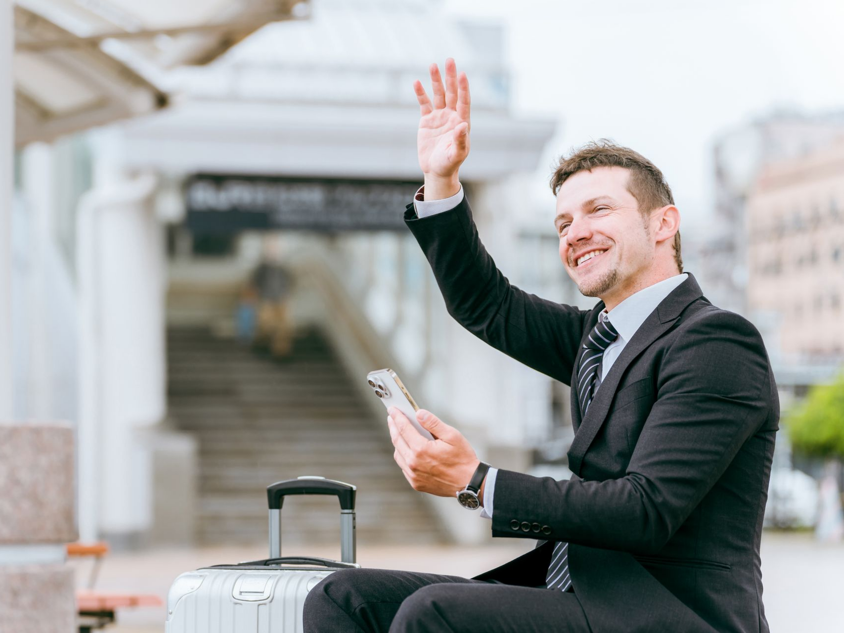 Un homme en costume, assis sur des bagages, fait signe de la main tout en tenant un téléphone.
