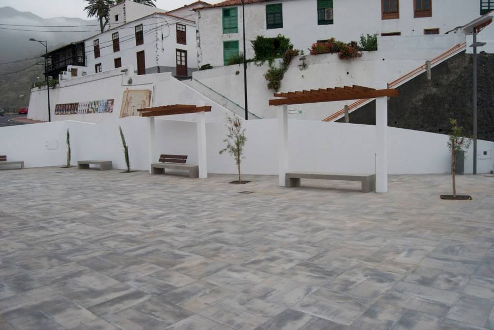 Edificios blancos en la ladera de una colina sobre una plaza empedrada con bancos y un pequeño árbol.