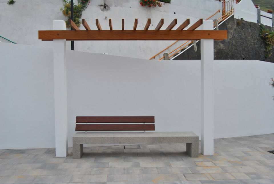 Banco exterior bajo una pérgola de madera, junto a una pared blanca, en un patio pavimentado.