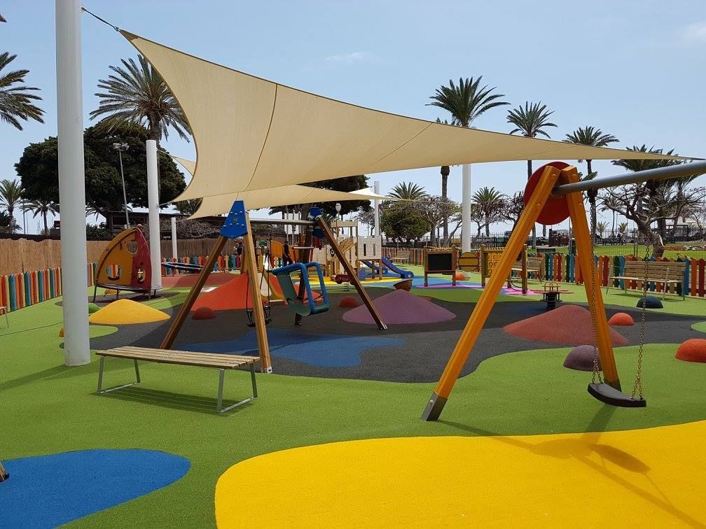 Parque infantil con coloridas velas de sombra, columpios y estructuras para trepar sobre un brillante suelo de goma.