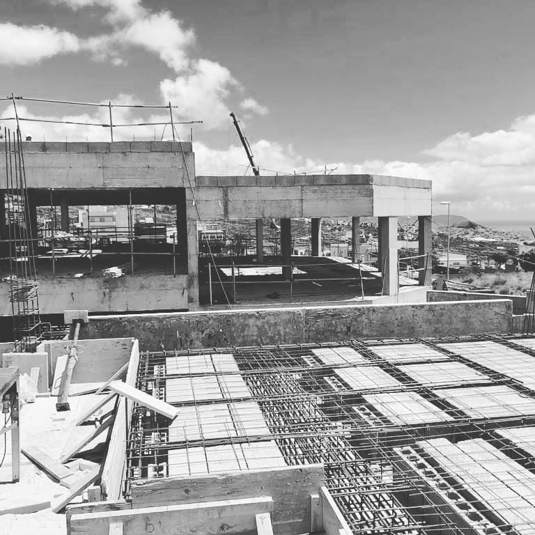 Obra en construcción con una estructura de hormigón armado, una grúa y una rejilla de barras de refuerzo en primer plano bajo un cielo nublado.